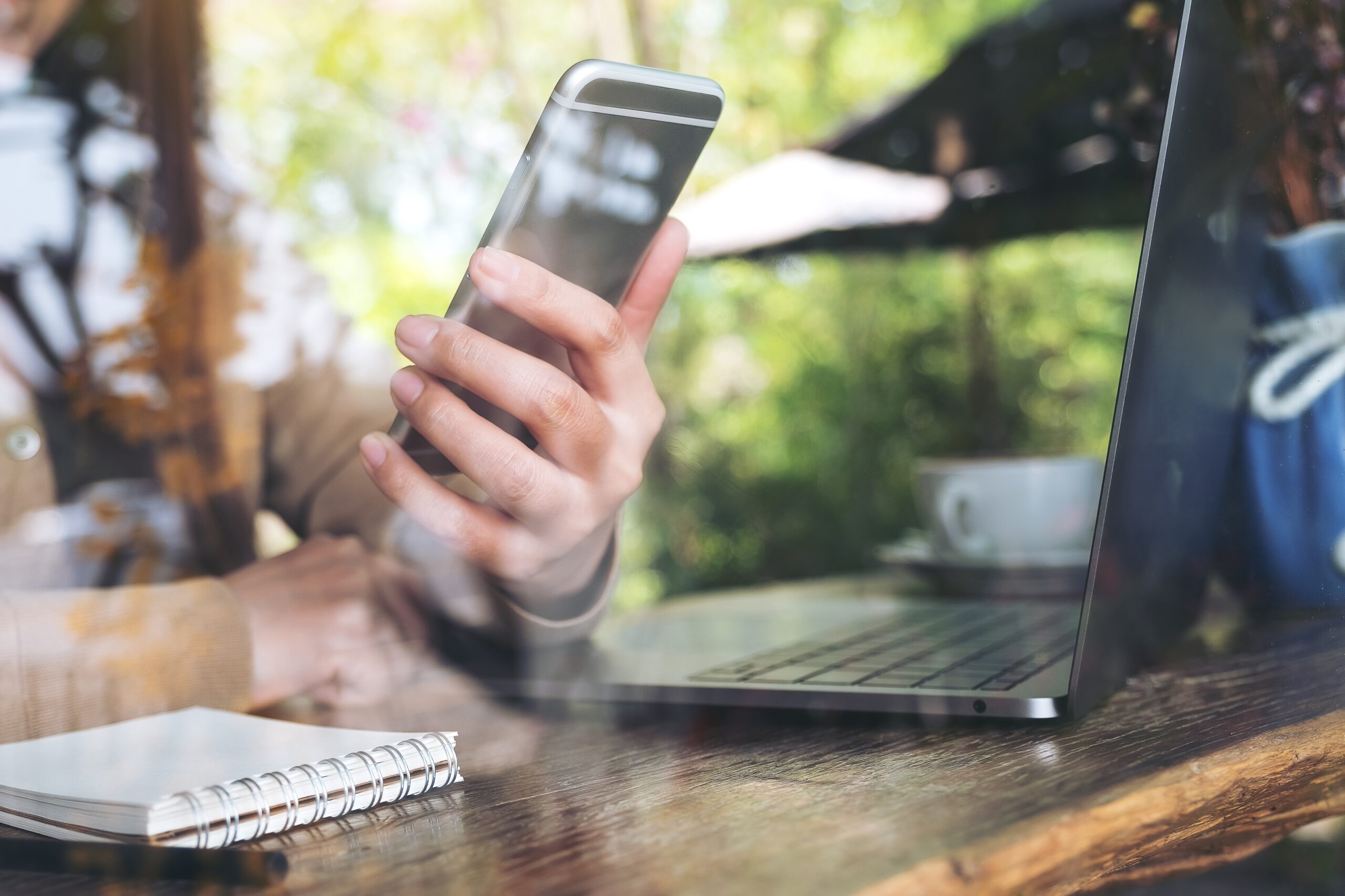 Closeup image of woman's hand holding and looking at smart phone while using laptop in cafe